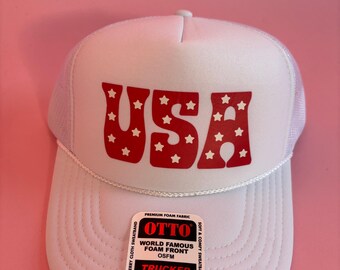 May include: White trucker hat with the word "USA" in red, star-filled letters. The hat has a white mesh back and a curved brim. The hat has a label that says "OTTO" and "TRUCKER".