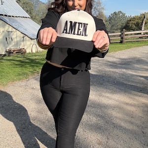 May include: A baseball cap with a white crown and a black brim. The word "AMEN" is printed in bold, black letters across the front of the cap. The cap is held up in front of a person.