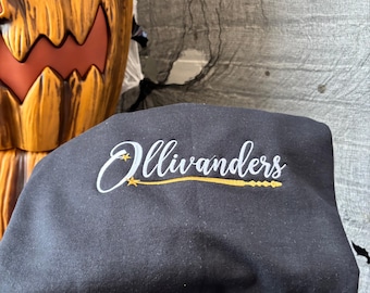 May include: Black sweatshirt with the text "Ollivanders" in white script and a gold wand design. The sweatshirt is folded, with a carved pumpkin and gray fabric in the background.