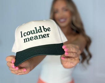May include: A white and dark green baseball cap with the text "I could be meaner" embroidered on the front. The cap has a curved brim and a classic trucker hat design.