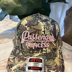May include: A camouflage print baseball cap with the text "Passenger PRINCESS" in pink script. The cap has a curved brim and a tag that reads "OTTO CLASSIC FIT OSFM CAMO". The hat is designed for maximum comfort and has a sporty style.
