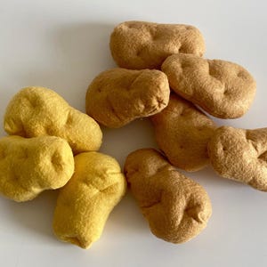 Op de afbeelding: Een verzameling handgemaakte vilten aardappelen in tinten geel en bruin. De aardappelen variëren in grootte en vorm, sommige zijn langwerpig en andere ronder. Het vilt heeft een getextureerd uiterlijk.