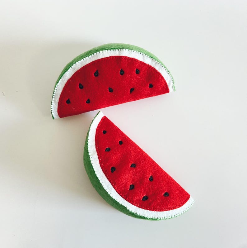 Pu&ograve; includere: Due fette di anguria in feltro. Il frutto rosso ha dettagli di semi neri e una buccia bianca, con un bordo esterno verde. Le fette sono disposte su uno sfondo bianco.