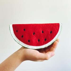 Pu&ograve; includere: Una fetta di anguria in feltro con interno rosso, semi neri e scorza bianca con cuciture verdi. L'oggetto artigianale &egrave; tenuto in mano su sfondo bianco. Un oggetto decorativo divertente.