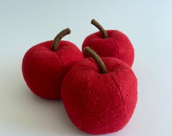 Può includere: Tre mele rosse di peluche con gambi marroni sono disposte su una superficie bianca. Le mele sono di un colore rosso vibrante e sembrano essere fatte di un materiale morbido. I gambi sono di colore marrone scuro.