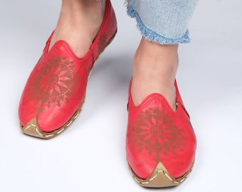 May include: A pair of red leather slip-on shoes with a decorative brown floral pattern on the vamp. The shoes have a pointed toe and a tan sole. The shoes are worn with light blue denim jeans.
