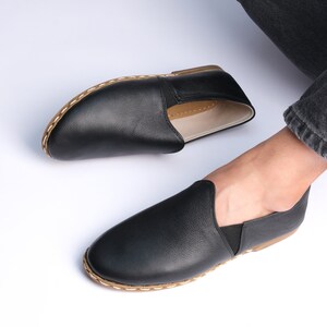 May include: A pair of black leather slip-on shoes with a brown stitched trim. The shoes have a simple design and are made of a soft, comfortable material.