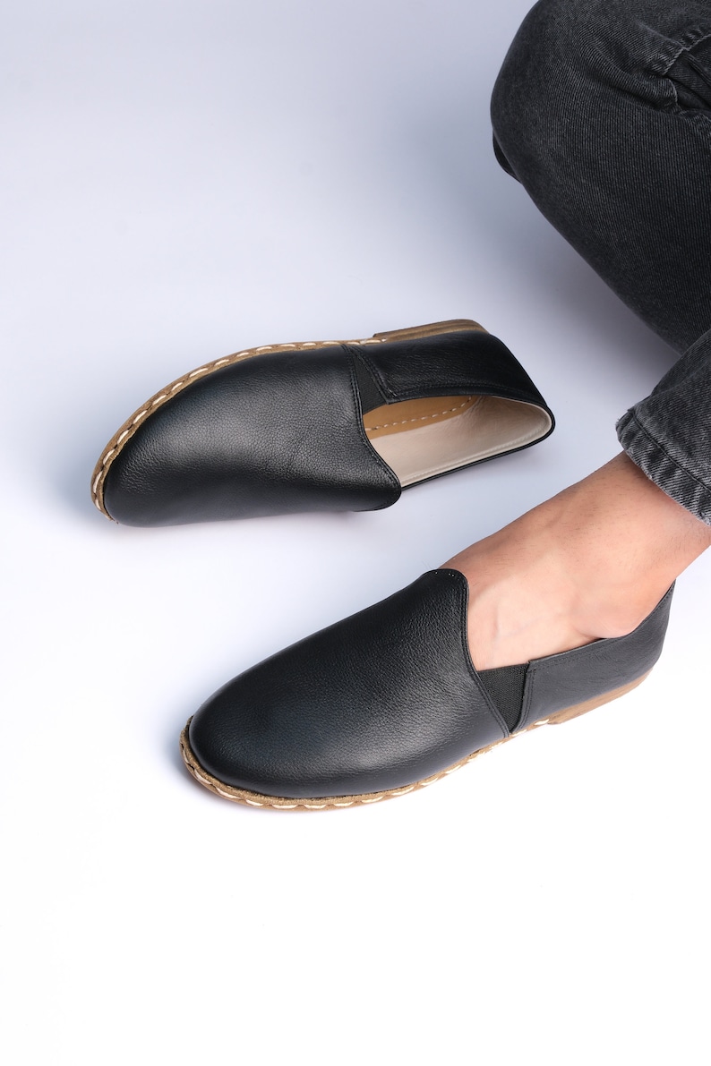 May include: A pair of black leather slip-on shoes with a brown stitched trim. The shoes have a simple design and are made of a soft, comfortable material.