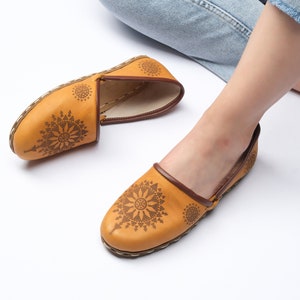 May include: A pair of tan leather slip-on shoes with intricate brown geometric designs. The shoes have a rounded toe and a flat sole.