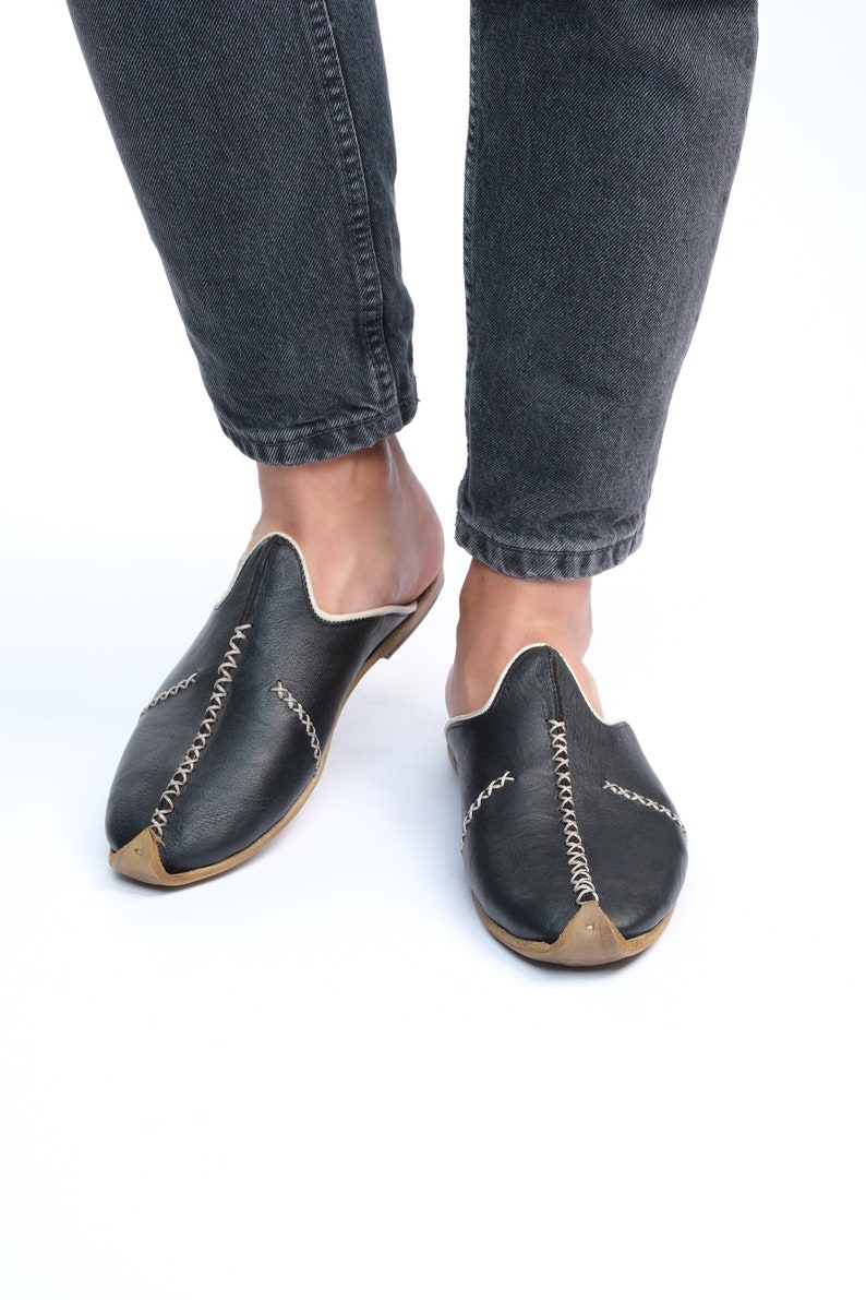 May include: A pair of black leather slippers with white stitching and a pointed toe. The slippers are worn by a person wearing dark denim jeans.