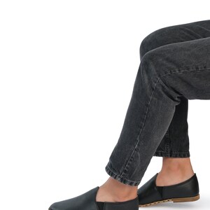 May include: A pair of black leather slip-on shoes with a brown stitched sole. The shoes are worn by a person wearing dark denim jeans.