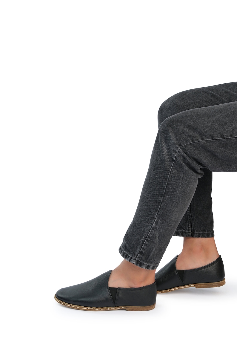 May include: A pair of black leather slip-on shoes with a brown stitched sole. The shoes are worn by a person wearing dark denim jeans.