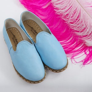 K&ouml;nnte beinhalten: Ein Paar hellblaue Lederschlappen mit braunem Steppsaum. Die Schuhe haben ein schlichtes Design und sind aus weichem Leder gefertigt.