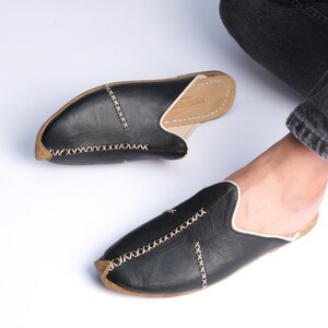 May include: A pair of black leather slippers with white stitching and a brown leather sole. The slippers have a pointed toe and a slip-on design.