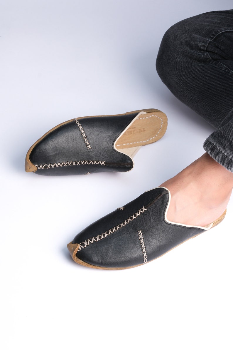 May include: A pair of black leather slippers with white stitching and a brown leather sole. The slippers have a pointed toe and a slip-on design.