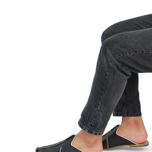 May include: A pair of black leather slip-on shoes with gold stitching. The shoes are worn by a person wearing dark denim jeans.