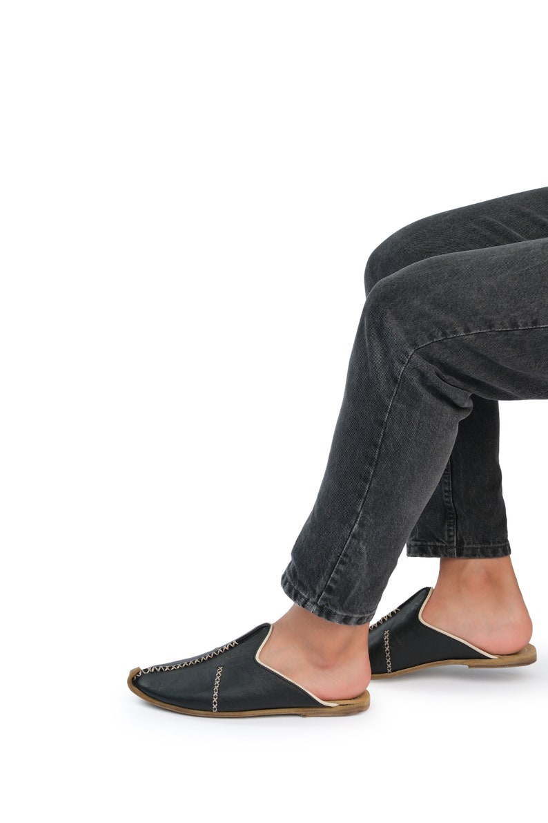 May include: A pair of black leather slip-on shoes with gold stitching. The shoes are worn by a person wearing dark denim jeans.