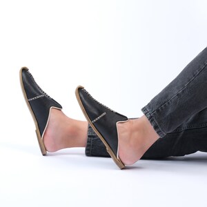May include: A pair of black leather slip-on shoes with a white stitch detail and a brown sole. The shoes are worn by a person with their legs crossed.