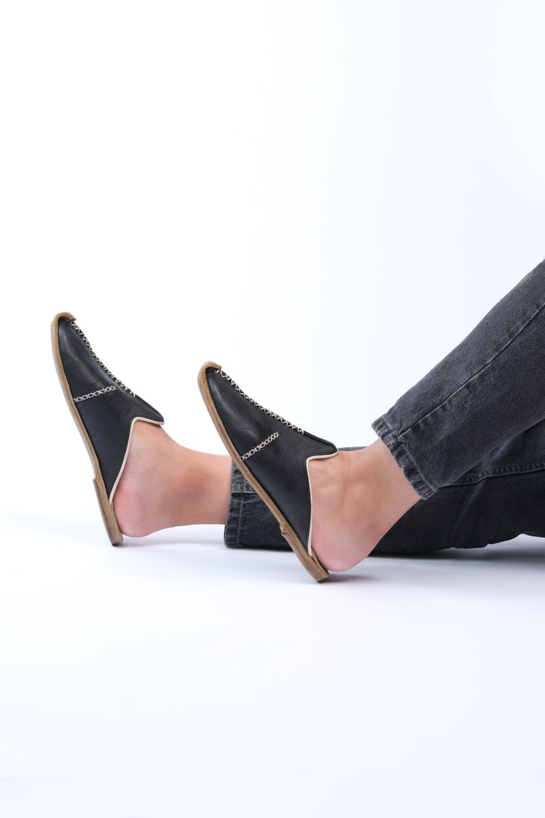 May include: A pair of black leather slip-on shoes with a white stitch detail and a brown sole. The shoes are worn by a person with their legs crossed.