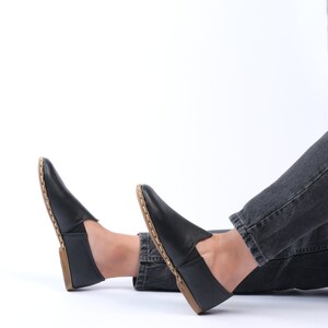May include: A pair of black leather slip-on shoes with a brown stitch detail around the edge. The shoes are worn by a person sitting on a white background.