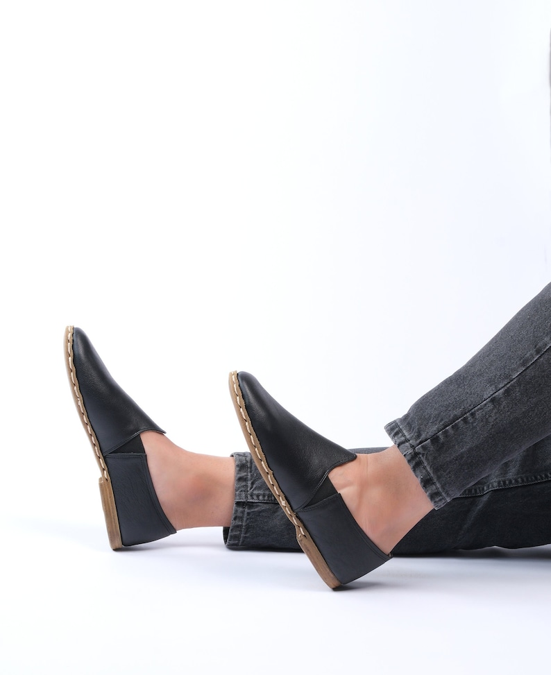 May include: A pair of black leather slip-on shoes with a brown stitch detail around the edge. The shoes are worn by a person sitting on a white background.