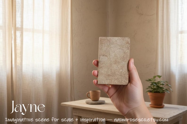 May include: A rectangular notebook with a textured, light beige cover featuring a marbled, organic pattern. The notebook is held upright, displaying its front cover. The text 'Jayne' is printed in white in the lower left corner of the image.