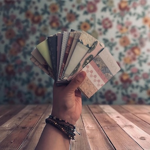 May include: A hand holding a fanned collection of small, patterned paper cards. The cards display various designs, including floral, geometric, and textured patterns in shades of pink, green, and neutral tones. The background features a floral wallpaper and a wooden surface.