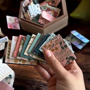 May include: A collection of miniature, patterned envelopes in various colors and designs, including floral and geometric patterns. Some envelopes are held in a hand, while others are in a wooden hexagonal box. The color palette includes pink, green, blue, and cream.