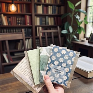May include: A set of four rectangular paper ephemera packs in clear plastic sleeves. The front pack features a blue and white polka dot pattern over vintage newspaper text with a label reading Hourglass Atrium. Other packs show green botanical prints.