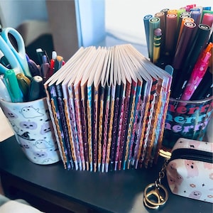 May include: A collection of small, hand-bound booklets with colorful stitched spines arranged in a row. The booklets are flanked by two cylindrical pen holders filled with various markers and pens, and a small pouch with a ghost pattern.