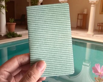 May include: A small, rectangular notebook with a light green, horizontally striped pattern. The notebook is held in front of a blurred background featuring a pool, architectural details, and pink flowers.