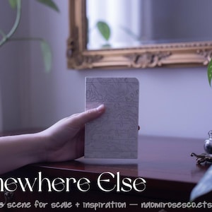 May include: A small rectangular notebook with a light gray cover featuring a detailed topographical map print. The notebook has rounded top corners and is held upright. The text "Somewhere Else" is printed in white at the bottom of the image.