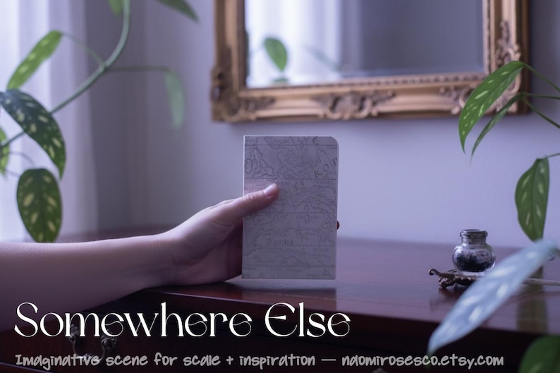May include: A small rectangular notebook with a light gray cover featuring a detailed topographical map print. The notebook has rounded top corners and is held upright. The text "Somewhere Else" is printed in white at the bottom of the image.