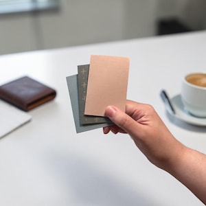 May include: A hand holds three small, rectangular notebooks. The top notebook is a light peach color, while the middle one is gray with a cross pattern, and the bottom one is a darker gray. A brown leather wallet and a cup of coffee are in the background.