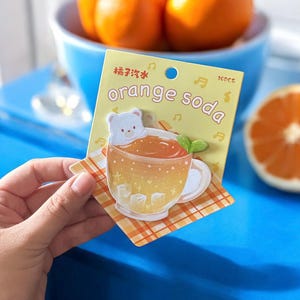 May include: A sticker featuring a cartoon illustration of orange soda in a teacup with a bear on top. The sticker has the words "orange soda" and other text in a yellow background. The sticker is on a checkered orange and white surface.