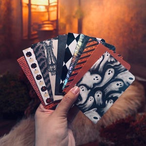 May include: A fan of rectangular cards featuring various Halloween-themed designs, including white ghosts on a black background, orange and black stripes, a black and white diamond pattern, a chandelier illustration, and small pumpkins on a red base.