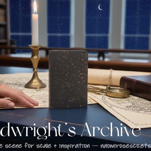 May include: A dark gray rectangular book featuring gold celestial illustrations of stars, moons, and constellations on the cover. The book is positioned upright on a surface alongside a brass candle holder with a lit candle, a compass, and maps.