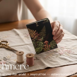 May include: A small rectangular notebook with a black cover featuring a botanical print of ferns and mushrooms in shades of green, orange, and brown. The notebook is displayed alongside a pair of gold-toned scissors and two small thread spools.
