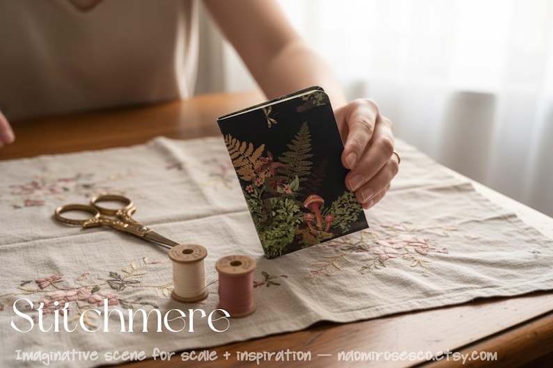 May include: A small rectangular notebook with a black cover featuring a botanical print of ferns and mushrooms in shades of green, orange, and brown. The notebook is displayed alongside a pair of gold-toned scissors and two small thread spools.