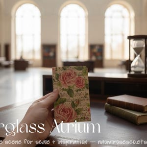 May include: A small rectangular card featuring a vintage-style floral pattern with pink roses and green leaves on a cream background with subtle text, held in front of a blurred library interior with an hourglass and two stacked books.
