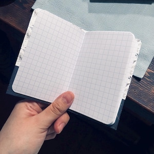 May include: Open notebook with grid paper, held by a hand. The notebook has a dark blue cover and decorative patterned edges. The pages are white with a gray grid.