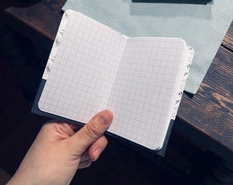 May include: Open notebook with grid paper, held by a hand. The notebook has a dark blue cover and decorative patterned edges. The pages are white with a gray grid.
