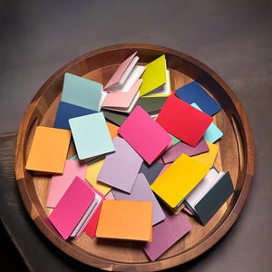 May include: A collection of small, rectangular, folded paper booklets in a variety of vibrant colors including pink, orange, yellow, blue, green, and purple, resting inside a circular wooden tray with a smooth, polished finish.