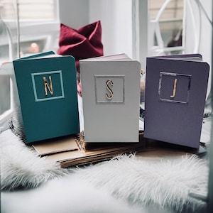 May include: Three personalized notebooks in teal, white, and purple. Each notebook features a gold initial within a square frame. The notebooks are displayed on a fluffy white surface, with a red fabric item in the background.