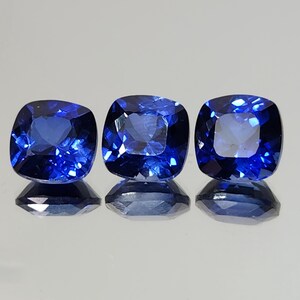 Pode incluir: São exibidas três safiras azuis escuras com corte almofada. Cada pedra preciosa tem uma forma quadrada com cantos arredondados e facetas que refletem a luz. As safiras são colocadas sobre um fundo claro, com seus reflexos visíveis abaixo.