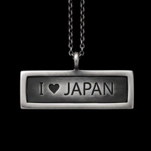 May include: A rectangular silver-toned pendant necklace featuring a brushed metal finish. The center of the pendant is engraved with the text I, a heart symbol, and the word JAPAN in capital letters. The pendant hangs from a matching silver chain.