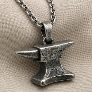 May include: A silver-toned metal pendant shaped like a blacksmith's anvil, featuring a textured, weathered finish. The pendant hangs from a matching silver-toned metal chain with rounded links, showcasing a sturdy and industrial aesthetic design.