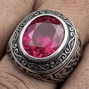 May include: A silver ring with an oval, faceted, ruby-red gemstone. The ring has an ornate, engraved design around the gemstone and on the band. The ring is a statement piece, suitable for special occasions.
