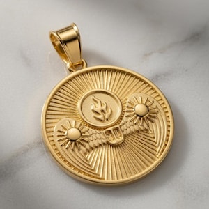 Puede incluir: Un colgante circular de oro con un emblema de llama central rodeado por dos soles estilizados con alas. El fondo del colgante tiene un patrón de rayos solares y cuenta con una reasa de oro pulido en la parte superior para una cadena.