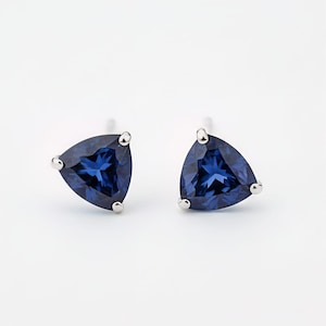 Puede incluir: Un par de pendientes de botón con piedras preciosas triangulares de color azul oscuro. Cada pendiente tiene un engaste plateado con tres pequeñas garras redondeadas. Los pendientes están sobre un fondo blanco.